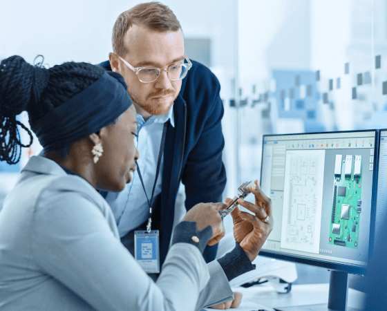 Two individuals discussing a mechanical component in front of a computer displaying technical diagrams. Two individuals discussing a mechanical component in front of a computer displaying technical diagrams.