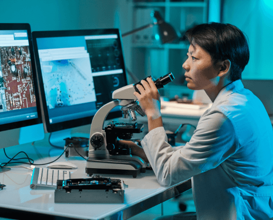 Person in a lab coat using a microscope at a desk with computer monitors showing technical images. Person in a lab coat using a microscope at a desk with computer monitors showing technical images.