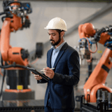 Man in a suit and hard hat holding a tablet in a factory with robotic arms.
