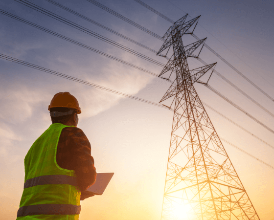 Utility worker in a hard hat and vest with a tablet near a transmission tower during sunset or sunrise. Utility worker in a hard hat and vest with a tablet near a transmission tower during sunset or sunrise.